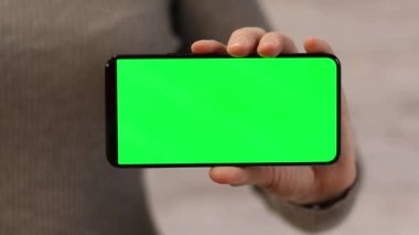 Elinde yeşil ekranlı bir telefon var. Elinde akıllı telefon tutan ince kazaklı bir kadın. Modern aletler. Cep telefonunun yakın çekimi. Krom anahtar, internette sörf yapmak, içeriği görmek
