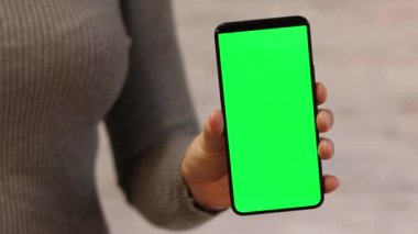 Elinde yeşil ekranlı bir telefon var. Elinde akıllı telefon tutan ince kazaklı bir kadın. Modern aletler. Cep telefonunun yakın çekimi. Krom anahtar, internette sörf yapmak, içeriği görmek