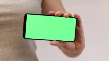 Elinde yeşil ekranlı bir telefon var. Elinde akıllı telefon tutan gri tişörtlü bir kadın. Modern aletler. Cep telefonunun yakın çekimi. Krom anahtar, internette sörf yapmak, içeriği görmek