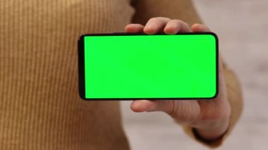 Elinde yeşil ekranlı bir telefon var. Elinde akıllı telefon tutan ince kazaklı bir kadın. Modern aletler. Cep telefonunun yakın çekimi. Krom anahtar, internette sörf yapmak, içeriği görmek