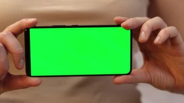 Elinde yeşil ekranlı bir telefon var. Yazlık tişörtlü bir kadın elinde akıllı telefon tutuyor. Modern aletler. Cep telefonunun yakın çekimi. Chromakey, internette sörf, reklam