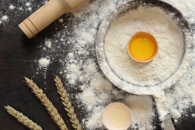 Pişirme süreci arka planı, üst görünüm. Çiğ yumurtalı un, hamuru sarmak için merdane, süzgeç ve buğday kulakları. Yemek pişirmek. Koyu masa üzerinde un yığını, hamur hazırlığı için eşyalar. Düz yatıyordu