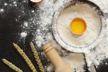 Pişirme süreci arka planı, üst görünüm. Çiğ yumurtalı un, hamuru sarmak için merdane, süzgeç ve buğday kulakları. Yemek pişirmek. Koyu masa üzerinde un yığını, hamur hazırlığı için eşyalar. Düz yatıyordu