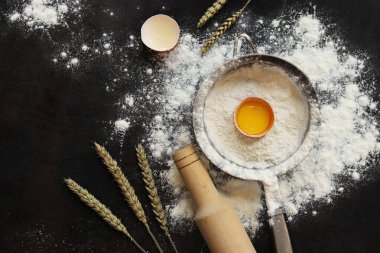 Pişirme süreci arka planı, üst görünüm. Çiğ yumurtalı un, hamuru sarmak için merdane, süzgeç ve buğday kulakları. Yemek pişirmek. Koyu masa üzerinde un yığını, hamur hazırlığı için eşyalar. Düz yatıyordu