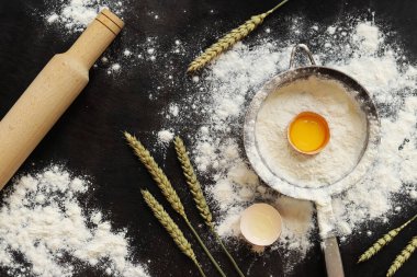 Pişirme süreci arka planı, üst görünüm. Çiğ yumurtalı un, hamuru sarmak için merdane, süzgeç ve buğday kulakları. Yemek pişirmek. Koyu masa üzerinde un yığını, hamur hazırlığı için eşyalar. Düz yatıyordu