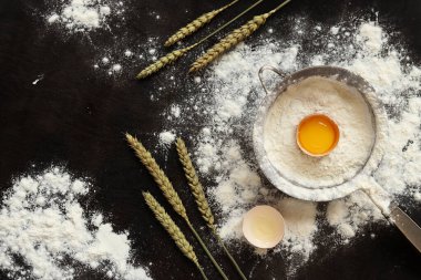 Çiğ yumurta, süzgeç ve buğday kulaklı un. Pişirme süreci arka planı, üst görünüm. Yemek pişirmek. Koyu masa üzerinde un yığını, hamur hazırlığı için eşyalar. Sağlıklı ev yemeği konsepti