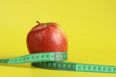 Elmanın etrafına sarılmış bir şerit. Metafor, yuvarlak şekil. Sarı arka planda olgun kırmızı elma ve mezura. Sağlıklı beslenme, diyet ve kilo kaybı kavramı. Vücut hacmi kontrolü
