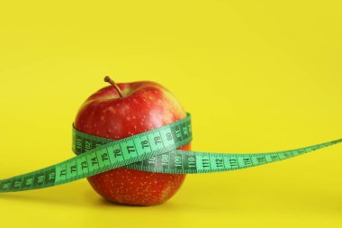 Elmanın etrafına sarılmış bir şerit. Metafor, yuvarlak şekil. Sarı arka planda olgun kırmızı elma ve mezura. Sağlıklı beslenme, diyet ve kilo kaybı kavramı. Vücut hacmi kontrolü