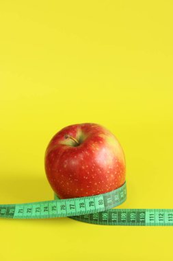 Elmanın etrafına sarılmış bir şerit. Metafor, yuvarlak şekil. Sarı arka planda olgun kırmızı elma ve mezura. Sağlıklı beslenme, diyet ve kilo kaybı kavramı. Vücut hacmi kontrolü