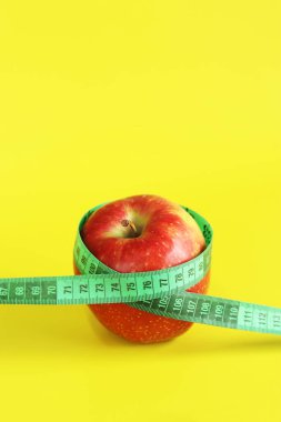 Elmanın etrafına sarılmış bir şerit. Metafor, yuvarlak şekil. Sarı arka planda olgun kırmızı elma ve mezura. Sağlıklı beslenme, diyet ve kilo kaybı kavramı. Vücut hacmi kontrolü