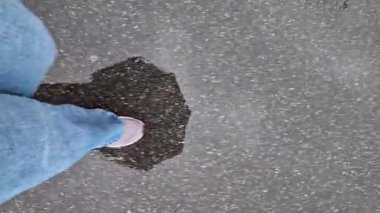 Islak asfalt üzerinde yürüyen insan ayaklarının yakın çekimi. Yağmurlu havada yürüyorum. Yürüyüş. Geniş mavi kotlu ve spor ayakkabılı kadın bacakları sokakta yürüyor, ilk bakışta. Yağmurda yürüyorum