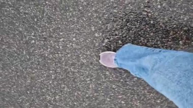 Islak asfalt üzerinde yürüyen insan ayaklarının yakın çekimi. Yağmurlu havada yürüyorum. Yürüyüş. Geniş mavi kotlu ve spor ayakkabılı kadın bacakları sokakta yürüyor, ilk bakışta. Yağmurda yürüyorum