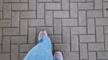 Kiremit kaplı bir yolda yürüyen insan bacaklarının yakın çekimi. Yağmurlu havada yürüyorum. Yürüyüş. Geniş mavi kot ve spor ayakkabılı kadın bacakları sokakta yürüyor, ilk bakışta görülüyor. Yağmurda yürüyorum