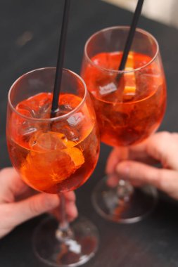 Kızların elinde Aperol Spritz olan iki bardak. Soğuk, ferahlatıcı alkollü kokteyl Aperol Spritz. Seçici odaklı kokteyller. Pipetle alkol içeceği. Geleneksel İtalyan içeceği