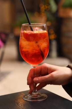 Elinde Aperol Spritz olan bir kadın. Soğuk, ferahlatıcı alkollü kokteyl Aperol Spritz. Bulanık bir arka planda kokteyl yakın plan. Pipetle alkol içeceği. Geleneksel İtalyan içeceği
