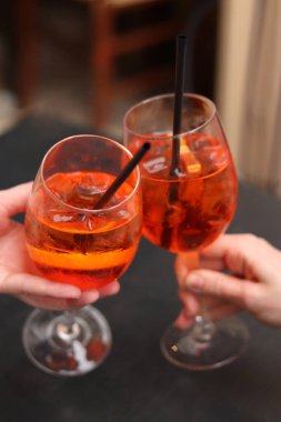 Kızların elinde Aperol Spritz olan iki bardak. Soğuk, ferahlatıcı alkollü kokteyl Aperol Spritz. Seçici odaklı kokteyller. Pipetle alkol içeceği. Geleneksel İtalyan içeceği