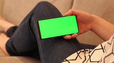 Yeşil ekran telefon. Akıllı telefondan bir şey izleyen bir kız. Yeşil ekran telefon. Evdeki kanepede oturan genç bir kadın elinde yeşil akıllı telefon ekran modeliyle. Krom anahtarlı cep telefonu. İnternet
