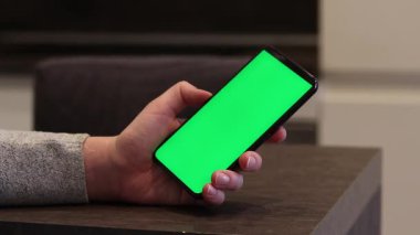 Elinde telefon olan bir kadın eli, yeşil ekran. İnternet araştırması. Modern aletler. Yeşil ekranlı akıllı telefon. Mutfakta oturan telefona bakan kız.