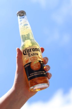 Kyiv, Ukrayna - 12 Mayıs 2025: Bir kadının elinde gökyüzüne karşı fazladan bira. Corona hafif bir bira. Şişe bira. Yakın planda meşrubat. Bir şişe Corona birası, arka ışıklandırma. Rahatlama