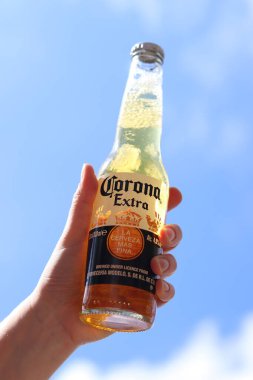 Kyiv, Ukrayna - 12 Mayıs 2025: Bir kadının elinde gökyüzüne karşı fazladan bira. Corona hafif bir bira. Şişe bira. Yakın planda meşrubat. Bir şişe Corona birası, arka ışıklandırma. Rahatlama