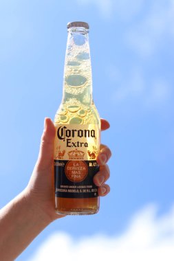 Kyiv, Ukrayna - 12 Mayıs 2025: Bir kadının elinde gökyüzüne karşı fazladan bira. Corona hafif bir bira. Şişe bira. Yakın planda meşrubat. Bir şişe Corona birası, arka ışıklandırma. Rahatlama