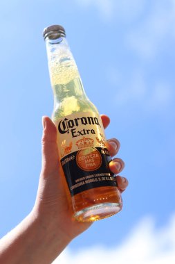 Kyiv, Ukrayna - 12 Mayıs 2025: Bir kadının elinde gökyüzüne karşı fazladan bira. Corona hafif bir bira. Şişe bira. Yakın planda meşrubat. Bir şişe Corona birası, arka ışıklandırma. Rahatlama