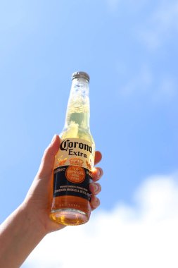 Kyiv, Ukrayna - 12 Mayıs 2025: Bir kadının elinde gökyüzüne karşı fazladan bira. Corona hafif bir bira. Şişe bira. Yakın planda meşrubat. Bir şişe Corona birası, arka ışıklandırma. Rahatlama