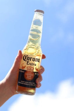 Kyiv, Ukrayna - 12 Mayıs 2025: Bir kadının elinde gökyüzüne karşı fazladan bira. Corona hafif bir bira. Şişe bira. Yakın planda meşrubat. Bir şişe Corona birası, arka ışıklandırma. Rahatlama