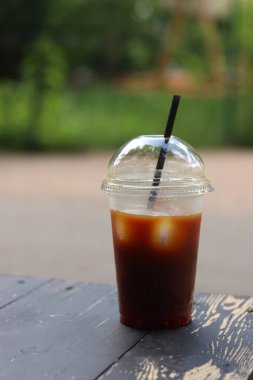 Tek kullanımlık kahve, ahşap masada, yakın plan. Arka planda bulanık park olan buzlu kahve. Plastik bardakta samanla birlikte soğuk kahve. Paket içki.