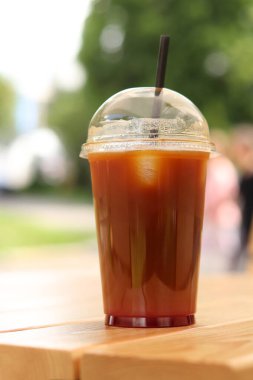 Tek kullanımlık kahve, ahşap masada, yakın plan. Arka planda bulanık park olan buzlu kahve. Plastik bardakta samanla birlikte soğuk kahve. Paket içki.