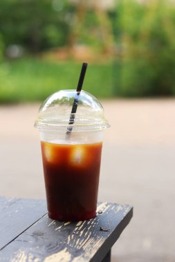 Tek kullanımlık kahve, ahşap masada, yakın plan. Arka planda bulanık park olan buzlu kahve. Plastik bardakta samanla birlikte soğuk kahve. Paket içki.