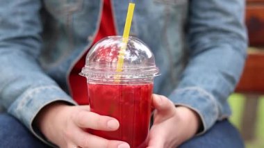 Elinde limonata olan kot ceketli bir kız, paket içecek. Yazın bir parkın arka planına karşı ferahlatıcı bir içecek. Tek kullanımlık bardakta içkiyle kadın ellerine yakın çekim. Yaz