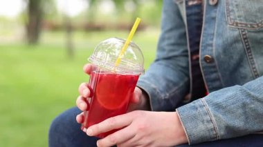 Elinde limonata olan kot ceketli bir kız, paket içecek. Yazın bir parkın arka planına karşı ferahlatıcı bir içecek. Tek kullanımlık bardakta içkiyle kadın ellerine yakın çekim. Yaz