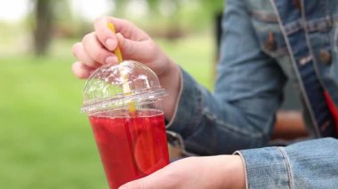 Tek kullanımlık plastik bardakta limonatalı bir kız, hazır içecek. Yazın bir parkın arka planına karşı ferahlatıcı bir içecek. Tek kullanımlık bardakta içkiyle kadın ellerine yakın çekim. Yaz