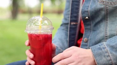 Tek kullanımlık plastik bardakta limonatalı bir kız, hazır içecek. Yazın bir parkın arka planına karşı ferahlatıcı bir içecek. Tek kullanımlık bardakta içkiyle kadın ellerine yakın çekim. Yaz