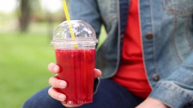 Elinde limonata olan kot ceketli bir kız, paket içecek. Yazın bir parkın arka planına karşı ferahlatıcı bir içecek. Tek kullanımlık bardakta içkiyle kadın ellerine yakın çekim. Yaz