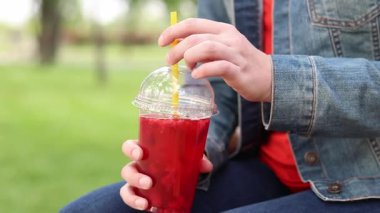 Tek kullanımlık plastik bardakta limonatalı bir kız, hazır içecek. Yazın bir parkın arka planına karşı ferahlatıcı bir içecek. Tek kullanımlık bardakta içkiyle kadın ellerine yakın çekim. Yaz