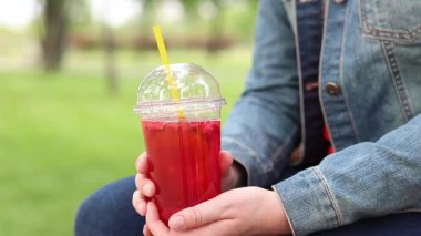 Tek kullanımlık plastik bardakta limonatalı bir kız, hazır içecek. Yazın bir parkın arka planına karşı ferahlatıcı bir içecek. Tek kullanımlık bardakta içkiyle kadın ellerine yakın çekim. Yaz
