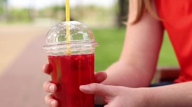 Tek kullanımlık bardakta limonata ile kadın ellerine yakın çekim. Yaz içeceği. Parlak tişörtlü kız parkta oturuyor. Yaz ve eğlence konsepti. Ahududu ve limonlu pembe limonata.