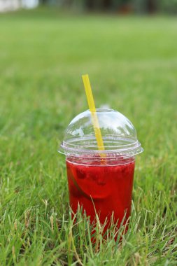 Tek kullanımlık plastik bardakta limonata çimlerin üzerinde duruyor. Yazın parkta serinletici bir içki. Ahududu ve limonlu pembe limonata. İçkiyi çimenlikteki tek kullanımlık bardakta yakın plan çek.