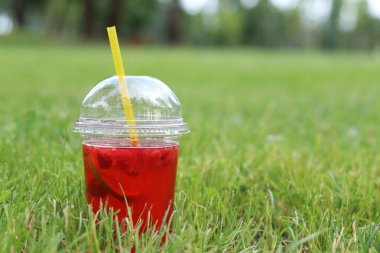 Tek kullanımlık plastik bardakta limonata çimlerin üzerinde duruyor. Yazın parkta serinletici bir içki. Ahududu ve limonlu pembe limonata. İçkiyi çimenlikteki tek kullanımlık bardakta yakın plan çek.