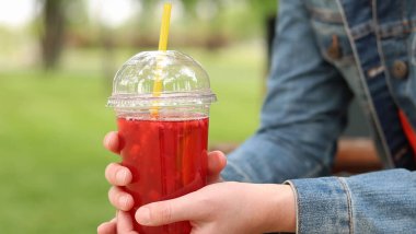 Elinde limonata olan kot ceketli bir kız, paket içecek. Yazın bir parkın arka planına karşı ferahlatıcı bir içecek. Tek kullanımlık bardakta içkiyle kadın ellerine yakın çekim. Yaz