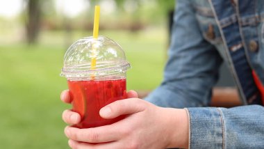 Elinde limonata olan kot ceketli bir kız, paket içecek. Yazın bir parkın arka planına karşı ferahlatıcı bir içecek. Tek kullanımlık bardakta içkiyle kadın ellerine yakın çekim. Yaz