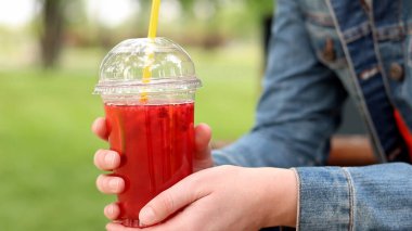 Elinde limonata olan kot ceketli bir kız, paket içecek. Yazın bir parkın arka planına karşı ferahlatıcı bir içecek. Tek kullanımlık bardakta içkiyle kadın ellerine yakın çekim. Yaz