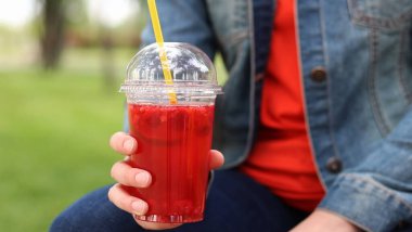 Elinde limonata olan kot ceketli bir kız, paket içecek. Yazın bir parkın arka planına karşı ferahlatıcı bir içecek. Tek kullanımlık bardakta içkiyle kadın ellerine yakın çekim. Yaz