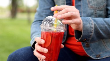 Elinde limonata olan kot ceketli bir kız, paket içecek. Yazın bir parkın arka planına karşı ferahlatıcı bir içecek. Tek kullanımlık bardakta içkiyle kadın ellerine yakın çekim. Yaz