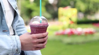 Dondurma ve böğürtlen şurubuyla kahve. Tek kullanımlık plastik bardakta samanla yaz içkisi içen kadın ellerine yakın çekim. Yaz günü ferahlatıcı bir içki. Kahveli meyveli milkshake. Parkta yürü.