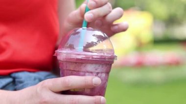 Dondurmalı meyveli milkshake. Tek kullanımlık plastik bardakta samanla yaz içkisi içen kadın ellerine yakın çekim. Yaz günü ferahlatıcı bir içki. Kahveli meyveli milkshake. Parkta yürü.