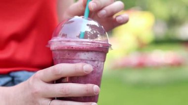 Dondurmalı meyveli milkshake. Tek kullanımlık plastik bardakta samanla yaz içkisi içen kadın ellerine yakın çekim. Yaz günü ferahlatıcı bir içki. Kahveli meyveli milkshake. Parkta yürü.