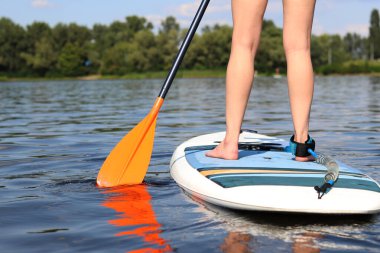 Kız bir sörf tahtasına biniyor, bacakları yakın plan. Aktif yaz tatili. Turist rotası için raket kiralama, su sporları. Kürek çeken bir kız sahilin yanındaki nehirde yüzüyor..